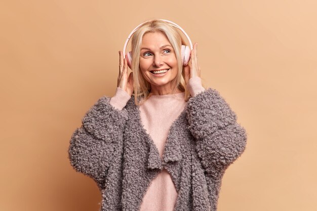 즐거운 미소를 지닌 세련된 금발의 유럽 여성은 스테레오 헤드폰을 착용하고 좋아하는 음악을 즐깁니다. 꿈꾸는 표정이 갈색 벽 위에 절연 모피 코트를 입습니다.
