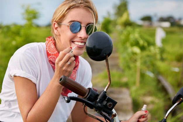 Fashionable blonde female biker wears shades paints lips with lipstick, looks in mirror of motorbike, cares about good appearance, likes transport and traveling in open air. Lifestyle and beauty