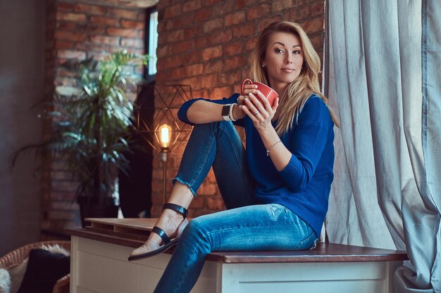 Fashionable blonde female holds cup of coffee while sitting on a table against a brick wall in a studio with a loft interior.