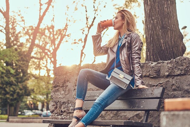 세련된 금발 여성은 야외에서 휴식을 취하며 도시 공원의 벤치에 앉아 테이크아웃 커피를 마십니다.