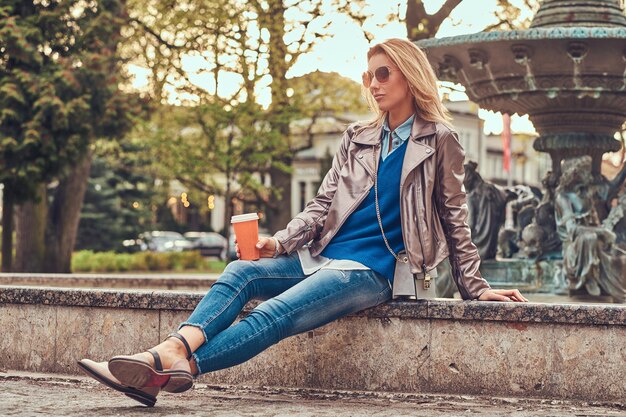 Fashionable blonde female relaxes outdoor, drinking takeaway coffee while sitting on the bench in city park.