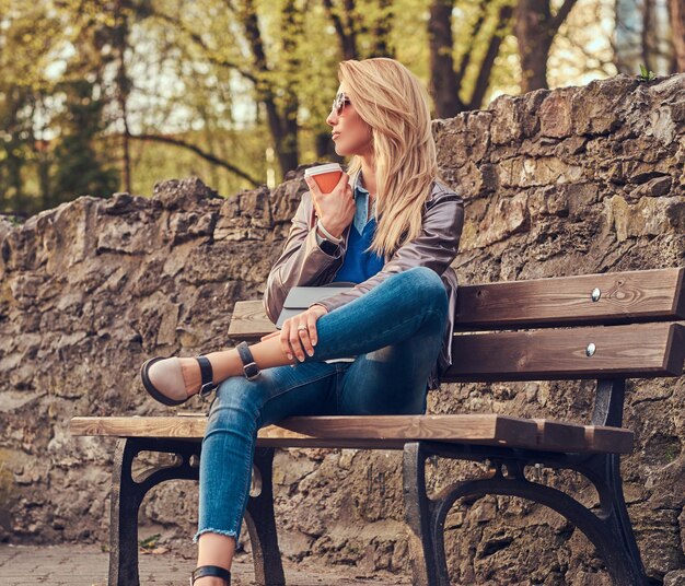 Модная блондинка отдыхает на свежем воздухе, пьет кофе на вынос, сидя на скамейке в городском парке.