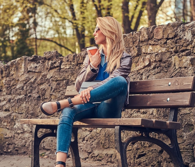Модная блондинка отдыхает на свежем воздухе, пьет кофе на вынос, сидя на скамейке в городском парке.