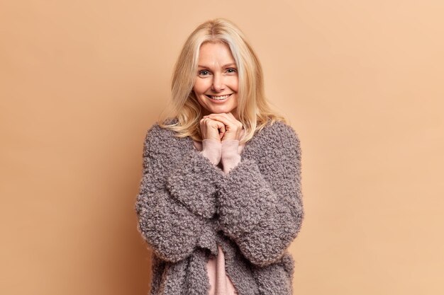 Fashionable blonde forty years old woman keeps hands under chin and smiles gently wears warm winter coat has minimal makeup poses against brown studio wall