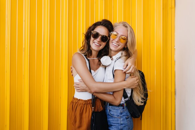 Fashionable blonde woman in headphones posing with brunette female friend on yellow background.