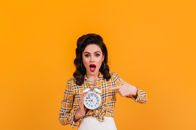 Fashionable brunette lady showing big clock. Studio shot of girl in vintage yellow attire.
