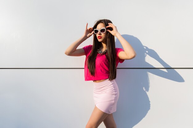 Fashionable brunette woman in summer clothes  and sunglasses posing over white urban wall.