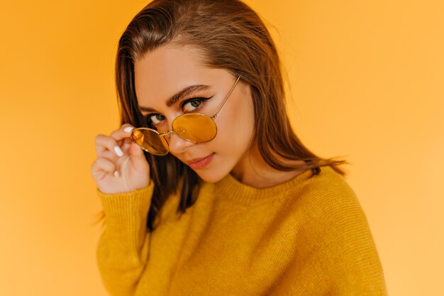 Fashionable caucasian girl looking through yellow sunglasses. Blissful female model in stylish autumn attire posing .