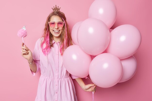 Fashionable cheerful young woman wears trendy sunglasses dress and crown holds fluffy pen and bunch of inflated balloons has good mood during birthday celebration glad to meet friends on party