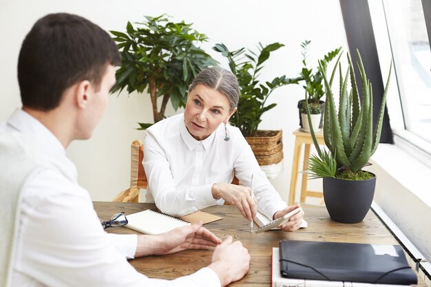 Fashionable confident dark haired elderly woman human recourses manager asking questions while having job interview with young man candidate who is applying for designer position. Selective focus