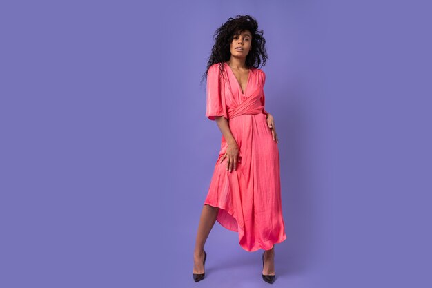 Модная уверенно женщина с вьющимися волосами позирует над фиолетовой стеной. Носить элегантное вечернее платье. Весенний модный образ. Полная длина.