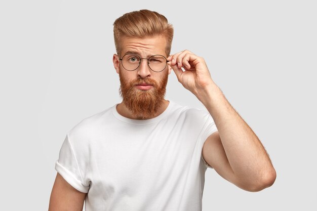 Fashionable cool man youngster with thick beard, blinks with eye, wears round spectacles