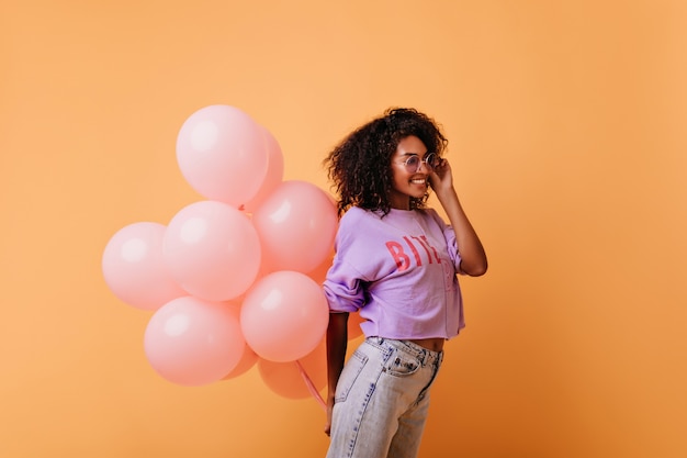 Fashionable curly woman in glasses holding pink helium balloons and laughing. pleased african birthday girl isolated on orange.