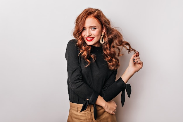 Fashionable curly woman in golden earrings standing on white wall. Adorable stylish girl laughing 