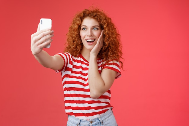세련된 귀여운 빨간 머리 곱슬머리 여성 여름 티셔츠 기록 비디오 소셜 미디어는 해외 럭셔리 열대 휴양지 여행에서 셀카를 들고 스마트폰 포즈를 취하는 어리석은 사진 빨간색 배경을 가지고 있습니다.