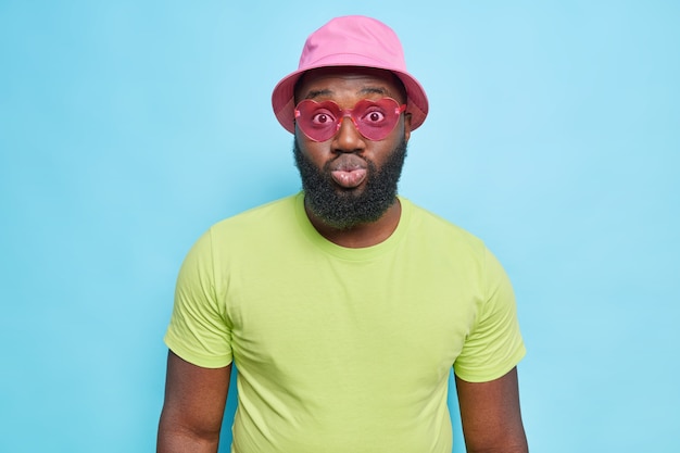 Fashionable dark skinned guy wears pink panama green t shirt and heart shaped sunglasses keeps lips rounded has romantic expression dressed in summer clothes