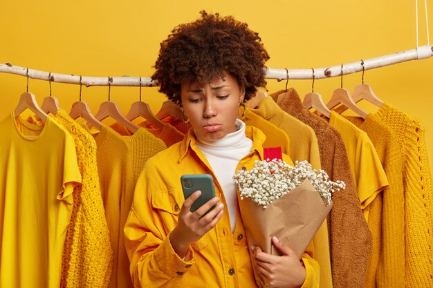 Fashionable displeased female customer looks upset, focused in smartphone, holds bouquet, poses against clothes in one tone hanging on rails, chooses outfit to wear