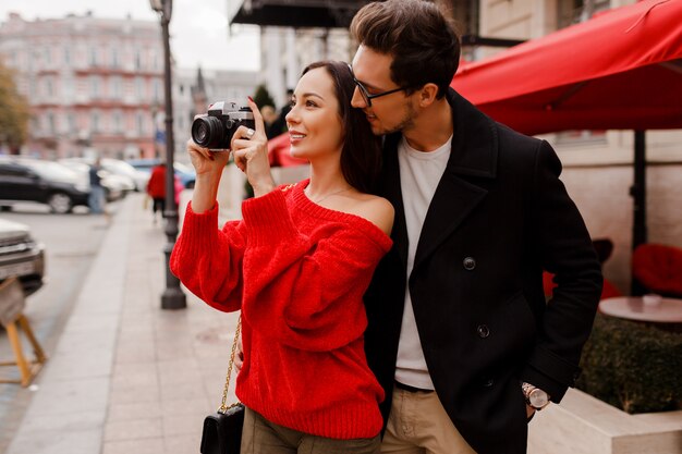 날짜 또는 휴일 동안 거리를 걷고 사랑에 유행 우아한 커플. 카메라로 사진을 만드는 빨간 스웨터에 갈색 머리 여자.