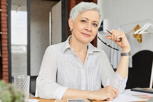 Fashionable elegant mature 50 year old female manager with gray pixie hair holding eyewear and  with confident smile, checking with financial papers, doing paperwork using calculator