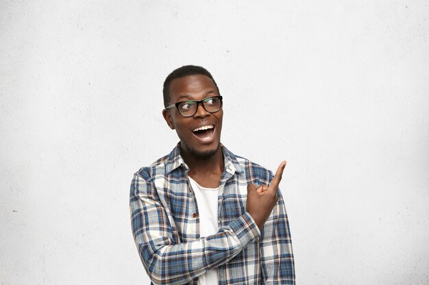 白い空白の壁で人差し指を指しているトレンディな眼鏡をかけているファッショナブルな感情的な若いアフリカ系アメリカ人男性