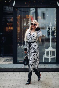 Fashionable european young woman in sunglasses walking outdoor at modern city exterior. attractive stylish blonde girl with handbag going at glass building background full shot