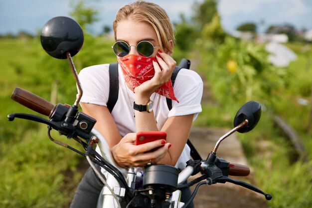 Fashionable female driver dressed casually, reads bikers blog on mobile phone, sits on motorbike, freshes fresh air outdoor, looks thoughtfully into distance. People, lifestyle and technology