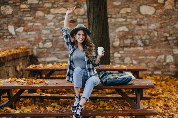 Free photo fashionable girl in short blue jeans sitting with cup of coffee in front of old building