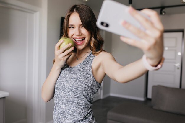 Fashionable girl taking picture if herself while eating apple. Indoor shot of fascinating lady using phone for selfie.