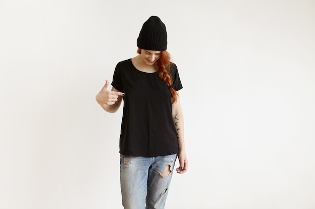fashionable girl with braid posing indoors in black hat and ragged jeans, pointing her finger at t-shirt