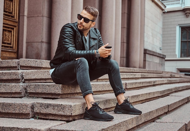 Free photo fashionable guy dressed in a black jacket and jeans holds a smartphone sitting on steps against an old building in europe.