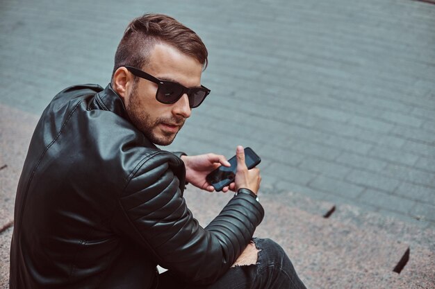 Fashionable guy dressed in a black jacket and jeans sitting on steps against an old building in Europe.