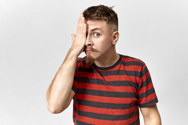 Fashionable hipster guy in stylish shirt with red and black stripes posing in gray studio, covering one eye, having his eyesight being examined. People, body language, signs, gestures and symbols