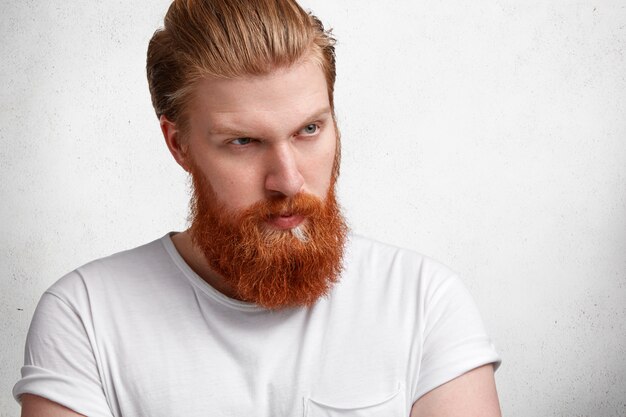 Fashionable hipster guy with stylish hairstyle, ginger thick beard and mustache, looks seriously and confidently into distance, contemplates about something