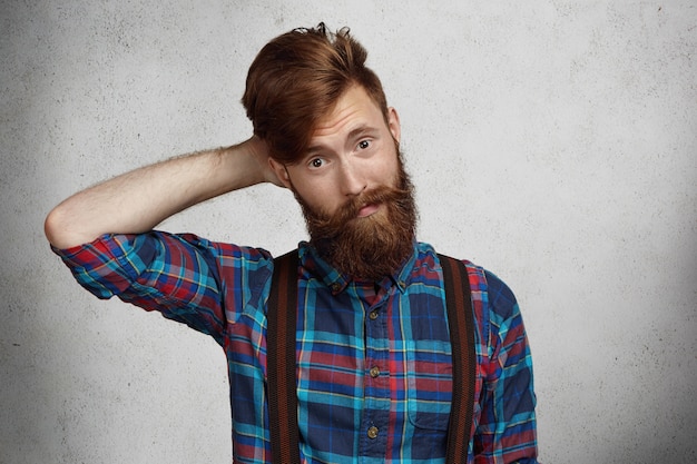 Fashionable hipster with thick beard dressed in trendy checkered shirt and suspenders looking puzzled and confused, holding hand behind his head.