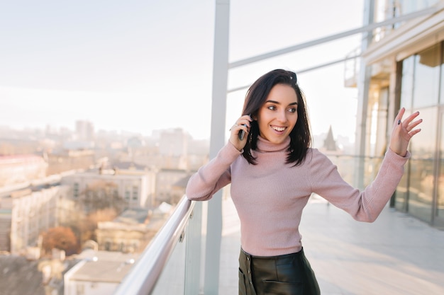 Fashionable joyful brunette young woman having fun on terrace on city view. Talking on phone, expressing true positive face emotions, happiness, excited, relax.