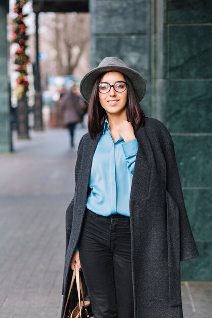 Fashionable joyful young businesswoman in black glasses walking on street in big city. Blue shirt, grey coat, hat, luxury clothes, elegant look, smiling to side, true emotions.