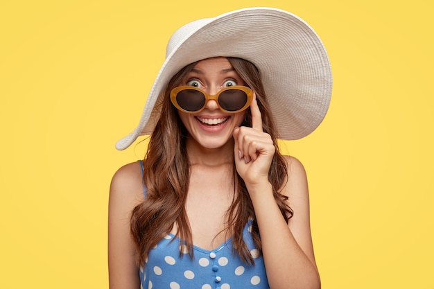 Fashionable lady with glad expression, wears white hat and sunglasses, finds hotel to stay during vacation, ready to go on beach, isolated over yellow wall. Tourism and summer time concept