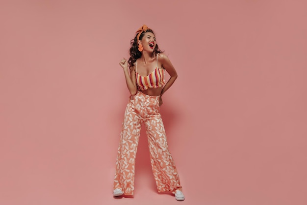 Free photo fashionable lady with tanned skin and red lips in modern earrings pineapple print pants and light sneakers laughing on pink backdrop