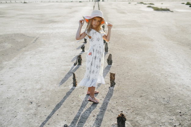 Free photo fashionable little girl model dressed in summer white dress is holding edges of the hat