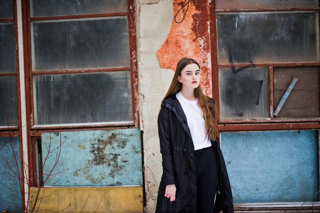 Fashionable long legs brunette model in long black cloak posed outdoor at winter day against old grunge wall with broken windows