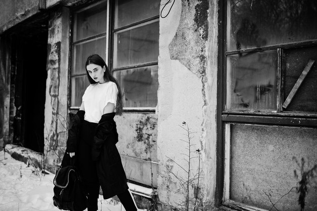 Fashionable long legs brunette model in long black cloak posed outdoor at winter day against old grunge wall with broken windows