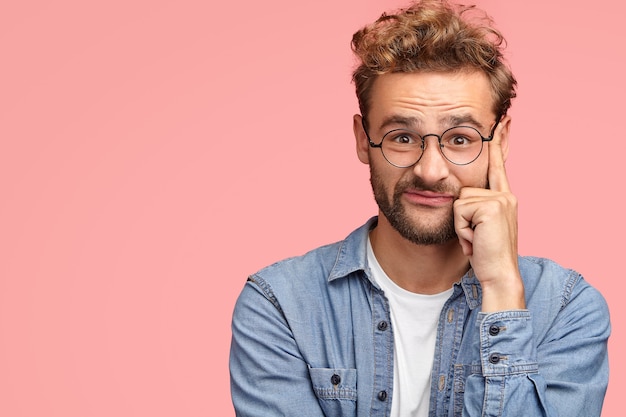 Fashionable male has curly hair and stubble touches temples and looks with seriousness, has puzzled facial expression, dressed in stylish denim shirt, poses over pink wall with copy space aside