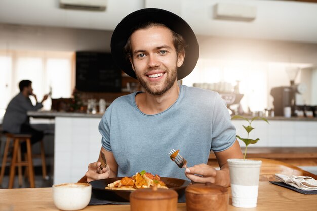 Fashionable male with beard appeasing hunger while dining alone at modern restaurant on sunny day, eating meal with knife and fork