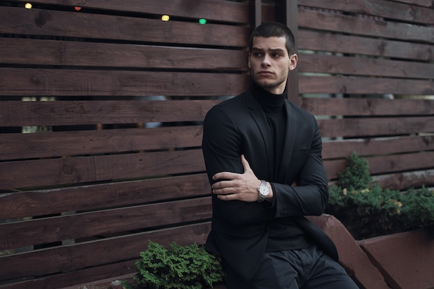 Fashionable man, wearing a suit, sitting against the wooden wall