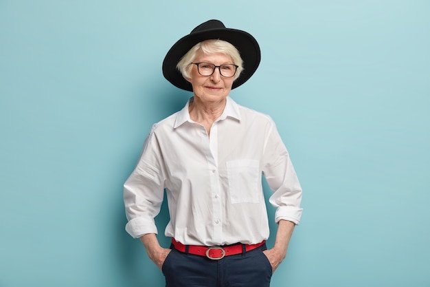 Fashionable mature woman wears stylish black hat, white shirt and trousers with red belt, keeps hands in pockets, has glad expression, isolated over blue wall. People, age and fashion concept