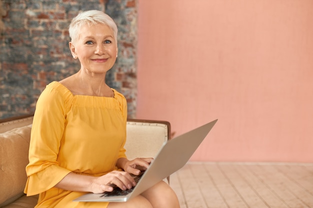 Free photo fashionable middle aged businesswoman checking email, sitting on couch with portable computer on her lap, keyboarding, using wireless high speed internet connection at home. people, age and technology