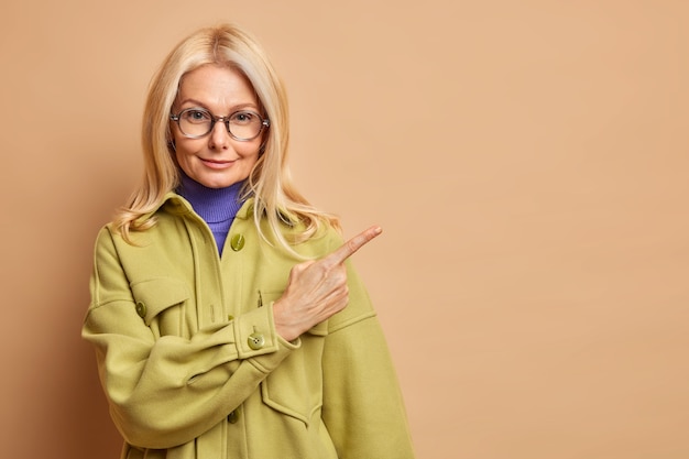 Модная женщина средних лет носит очки и зеленое осеннее пальто, указывая на пространство для копирования