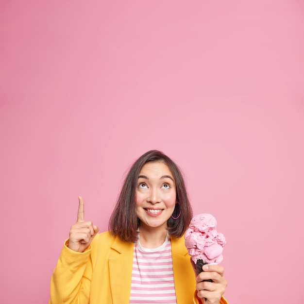 Fashionable millennial girl with short dark hair points overhead on blank copy space demonstrates something over pink wall eats yummy raspberry ice cream containing much calories