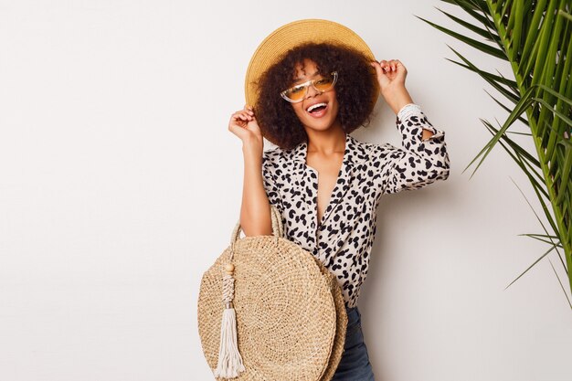 Fashionable mix race girl in trendy blouse and straw hat  standing over white wall. Perfect white  smile. Shopping mood.