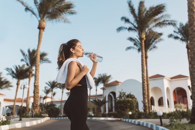 Fashionable pretty young woman in sportswear drinking water on street on palm trees, blue sky. Cheerful mood, closed eyes, relax, workout, fitness